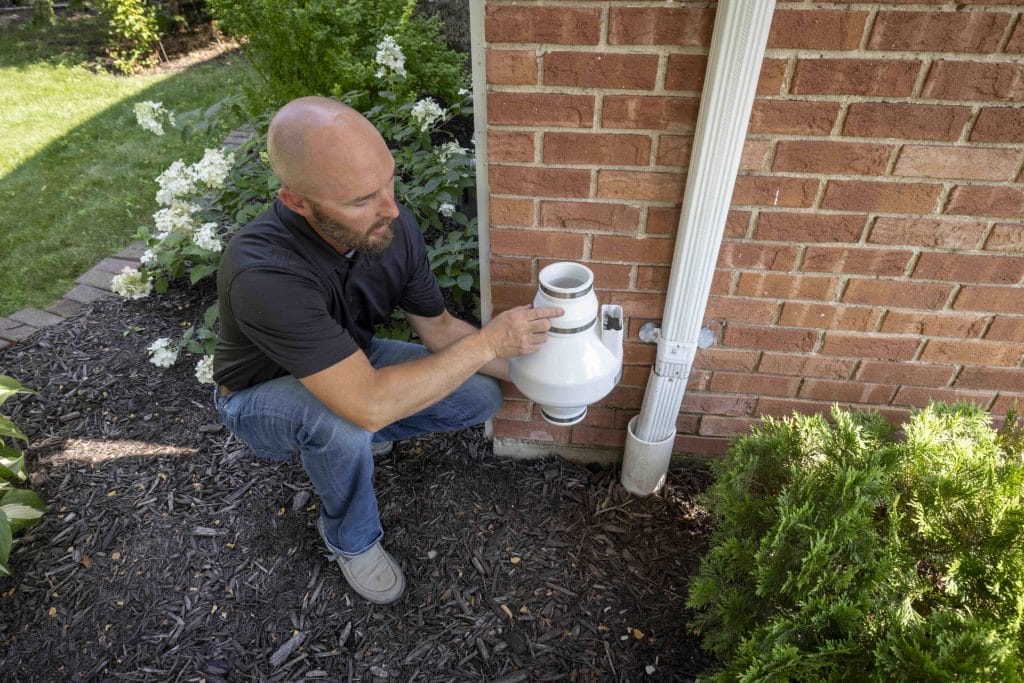 Man Inspecting Radon Mitigation System Outside Brick Home

