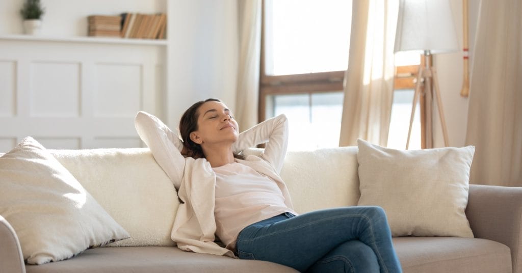 In a neutral living room, a woman leans back on a couch with her arms behind her head. Her eyes are closed.