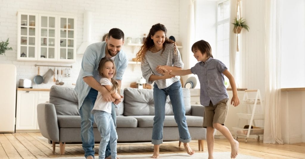 Two adults and two children stand and play together on a rug in a living room. The room is bright and airy.