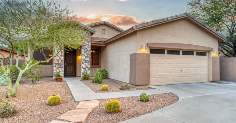 A southwestern-style home has stucco and stone walls. The front yard features barrel cacti, some other plants, and gravel.