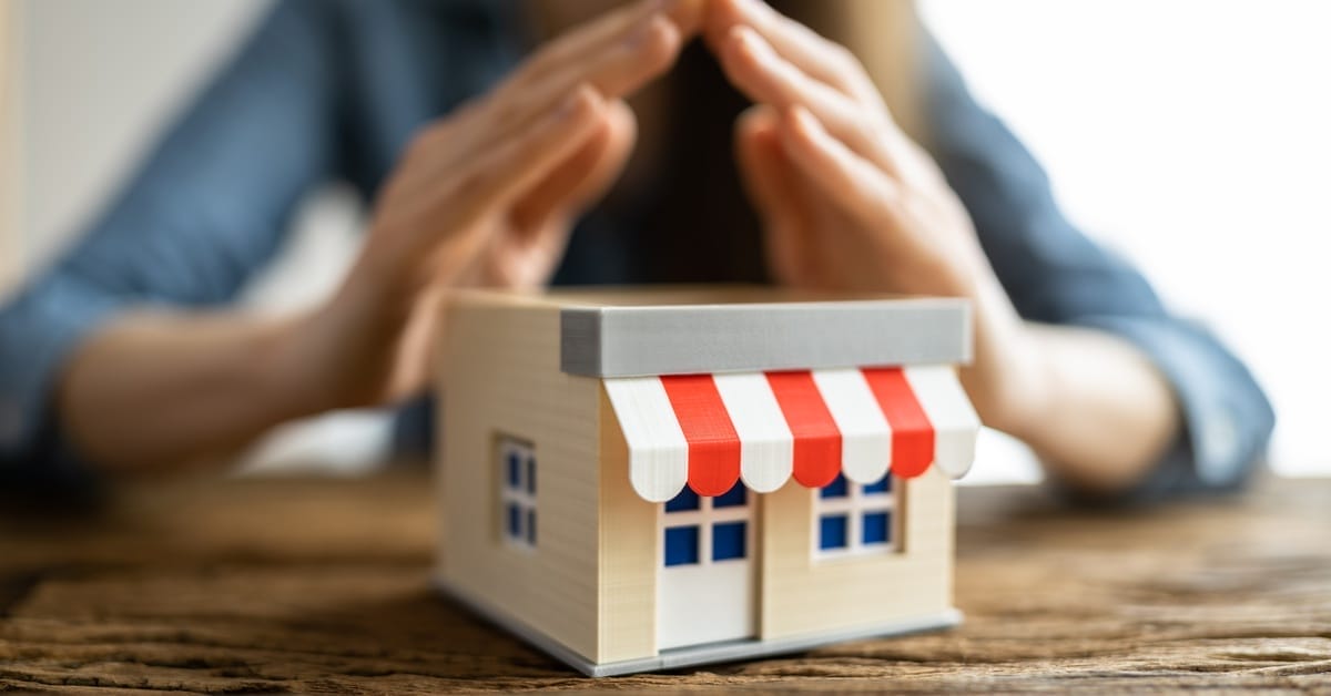 A miniature model of a small business with an awning is on a table. A person places their steepled hands over the model.