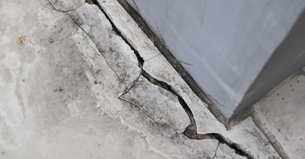 Downward view of a wide and long crack in a concrete floor next to a wall or pillar. The concrete is chipping away.