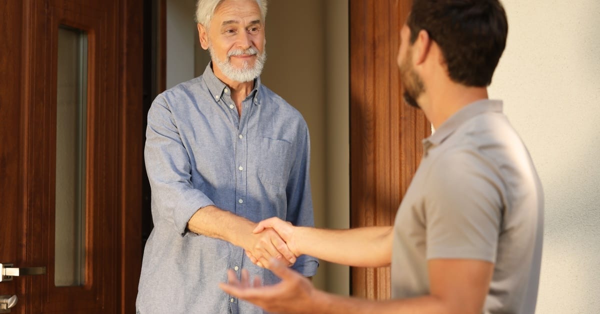 On a sunny day, a man stands in his home's doorway and shakes hands with another man who is standing just outside.